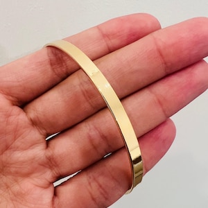 Può includere: Un braccialetto piatto color oro. Il braccialetto ha una superficie liscia e riflettente e un design semplice e minimalista. Il braccialetto è mostrato su uno sfondo neutro.