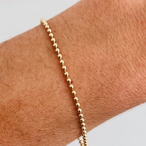 May include: A gold beaded bracelet. The bracelet is composed of small, round, gold beads strung together. The bracelet is shown on a wrist, with the beads catching the light.