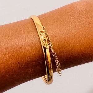 Puede incluir: Un brazalete dorado con un cierre de bisagra y una delicada cadena. El brazalete es liso y brillante, reflejando la luz. La cadena cuelga del cierre, agregando un elemento decorativo. La joya se lleva en el brazo de una persona.
