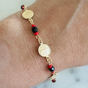 May include: Gold-toned bracelet featuring circular medallions and alternating red and black beads. The bracelet is connected by a delicate chain and is worn on a wrist. The design incorporates religious iconography.