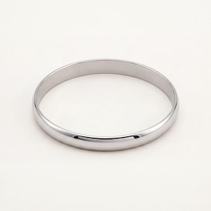 May include: A polished silver-tone bangle bracelet with a smooth, rounded exterior surface and a simple circular design. The metal has a reflective finish that catches the light, creating a clean and uniform appearance for this solid metal wrist accessory.