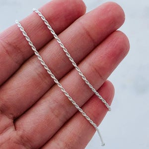 May include: A close-up of a silver-coloured rope chain. The chain is held in a hand, and the links are tightly woven together. The chain is a bright, shiny silver colour.