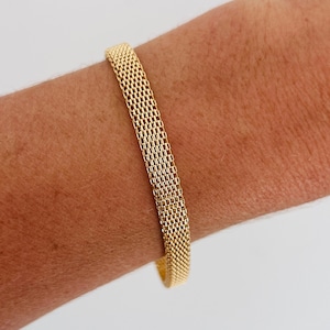 Op de afbeelding: Een goudkleurige mesh armband. De armband is gemaakt van een geweven metalen materiaal, waardoor een textuur ontstaat. De armband wordt om de arm van een persoon gedragen, wat het ontwerp en de stijl laat zien. De breedte van de armband is ongeveer 2 cm.