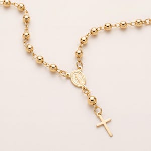 Può includere: Una collana rosario color oro caratterizzata da una serie di perline sferiche lucide collegate da sottili maglie metalliche. Il design include una medaglia centrale di forma ovale raffigurante una figura e un piccolo crocifisso semplice.