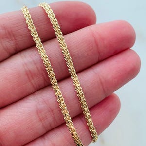 May include: Close-up of two delicate gold chains with a detailed, textured design. The chains are held in a hand, showcasing their intricate pattern and warm, golden color. The chains appear to be jewelry.