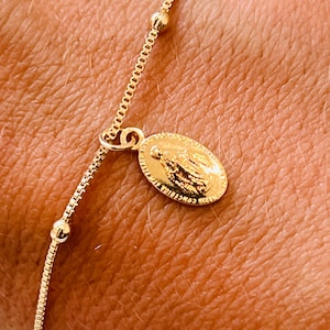 May include: A gold-colored bracelet featuring a delicate chain with small, round beads and a rectangular link chain. A small, oval pendant with a religious figure hangs from the chain. The bracelet is shown on a person's wrist.