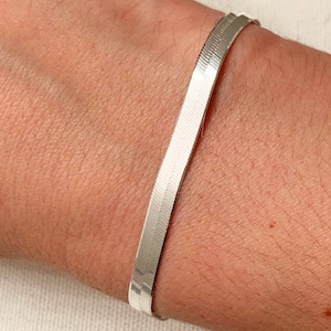 May include: A silver herringbone bracelet. The bracelet is made of small, flat, interlocking metal pieces, creating a smooth, flexible band. The bracelet is worn on a wrist, showcasing its sleek design and reflective surface.