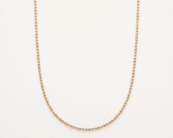 Könnte beinhalten: Eine goldfarbene Kugelkette, die aus einer durchgehenden Reihe kleiner, gleichmäßiger, kugelförmiger Perlen besteht, die zu einem zarten, flexiblen Strang verbunden sind. Die Kette ist in einer einfachen, symmetrischen U-Form angeordnet.