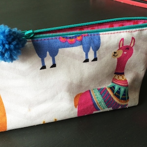 Puede incluir: Un estuche de lápices de lona blanca con un diseño de llama rosa, azul y naranja. La cremallera es turquesa y el tirador es un pompón azul.