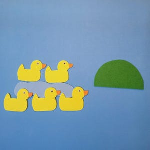 May include: A felt craft scene featuring five yellow duck cutouts with orange beaks and small black eyes. They are arranged on a light blue cutout representing water, with a green half-circle representing a hill, all against a blue background.