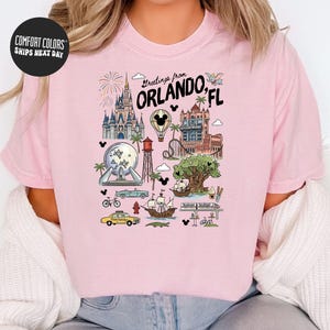 A pink t-shirt with a graphic design depicting various landmarks and attractions in Orlando, Florida, including Disney World, the Walt Disney World Resort, and other iconic sites.