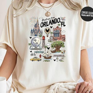 A person wearing a white t-shirt with a graphic design depicting various landmarks and attractions in Orlando, Florida.
