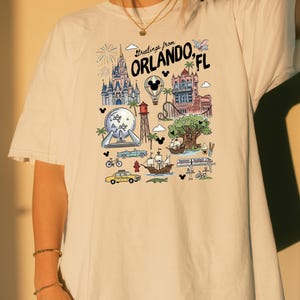 A person wearing a white t-shirt with a graphic design depicting various landmarks and attractions from Orlando, Florida.