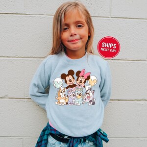 A young girl wearing a light blue sweatshirt with a cartoon character design, smiling at the camera.