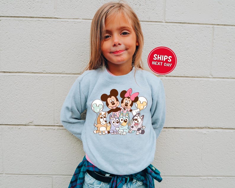 A young girl wearing a light blue sweatshirt with a cartoon character design, smiling at the camera.