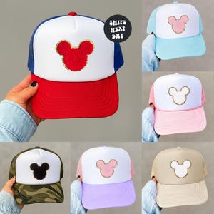 May include: A collection of trucker hats in various colors, each featuring a Mickey Mouse silhouette. The hats have a mesh back and a solid front panel. The hats are red, blue, pink, light blue, green camouflage, lavender, and tan.