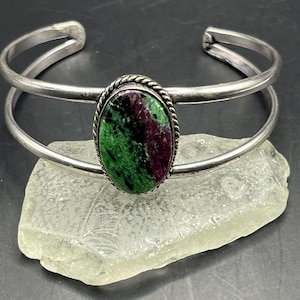 May include: A silver cuff bracelet featuring a double-band design and a central oval-shaped stone. The stone displays a mottled pattern of deep magenta, dark green, and black, surrounded by a twisted rope-style metal border frame.