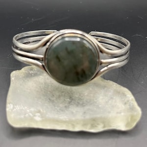 May include: A silver cuff bracelet with a large, round, dark green stone at the center. The bracelet has a simple, elegant design with three parallel bands on each side of the stone. The stone appears to be polished and smooth.
