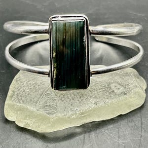 May include: A silver cuff bracelet featuring a rectangular, vertically striped dark green and black gemstone set in a polished metal frame. The band consists of two thin, smooth silver wires that meet at the central stone setting.