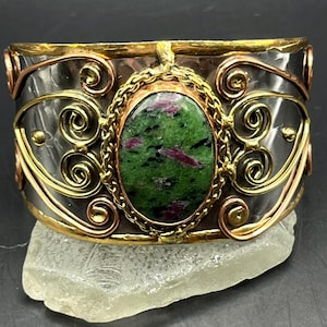 May include: A wide cuff bracelet featuring a central oval green and magenta zoisite stone set in a gold-toned braided bezel. The band is made of dark hammered metal with decorative gold and copper-toned wire swirls and scrollwork accents.