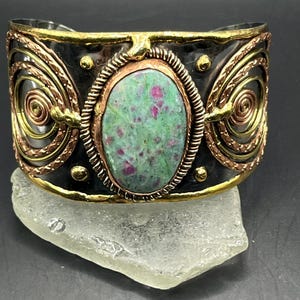 Peut inclure: Un bracelet manchette large orné d'une pierre ovale centrale verte et magenta, sertie dans une bordure en cuivre avec un fil torsadé. La bande en métal sombre est rehaussée de bords dorés, de petits clous et de spirales en cuivre et laiton.