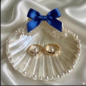 May include: A white seashell ring dish decorated with small white pearls along the edges and a vibrant blue satin ribbon bow at the top. Two gold-toned rings, one featuring a single clear stone, rest inside the shell on the pearled surface.