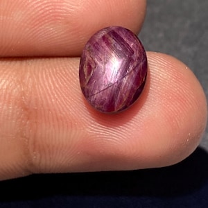 Könnte beinhalten: Ein ovaler, im Cabochon-Schliff gefertigter Edelstein in tiefem Magenta und Dunkelrot. Der Stein hat eine glatte, polierte Oberfläche mit sichtbaren inneren Einschlüssen und natürlichen Streifen, die ein strukturiertes Aussehen erzeugen.
