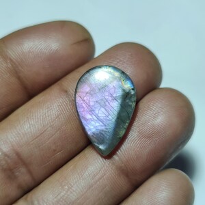 Puede incluir: Una piedra preciosa en forma de lágrima con una superficie pulida. La piedra muestra una mezcla de colores, incluyendo púrpura, azul y gris, con vetas internas. La piedra preciosa se sostiene en una mano, mostrando su tamaño y forma. Esta es una piedra preciosa para la fabricación de joyas.