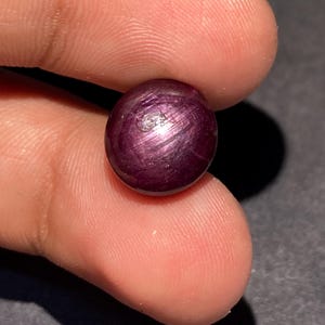 May include: A round, cabochon-cut star ruby gemstone with a deep, saturated purplish-red color. The surface displays a distinct, luminous six-rayed star effect, known as asterism, which shifts across the polished, smooth, and domed surface.