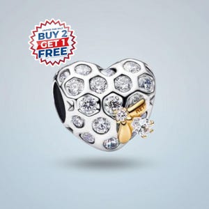 May include: A heart-shaped charm with a honeycomb design, featuring clear crystals and a gold-colored bee accent. The charm has a black opening on the side. A red, white, and blue sticker with the text "BUY 2 GET 1 FREE" is in the upper left corner.