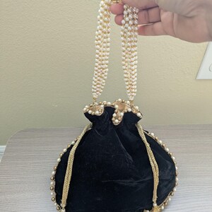 May include: Black velvet pouch with gold trim and pearl accents. The drawstring bag features a gold beaded trim and a gold and pearl handle. The bag is held by a hand.