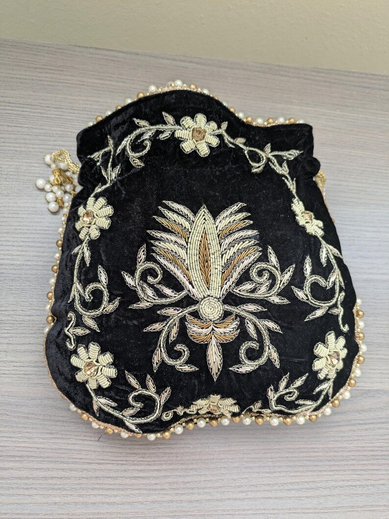 May include: Black velvet drawstring bag with elaborate gold and cream floral embroidery. The bag is trimmed with gold beads and faux pearls. A gold tassel with faux pearls hangs from the drawstring closure. The bag is a small, decorative accessory.