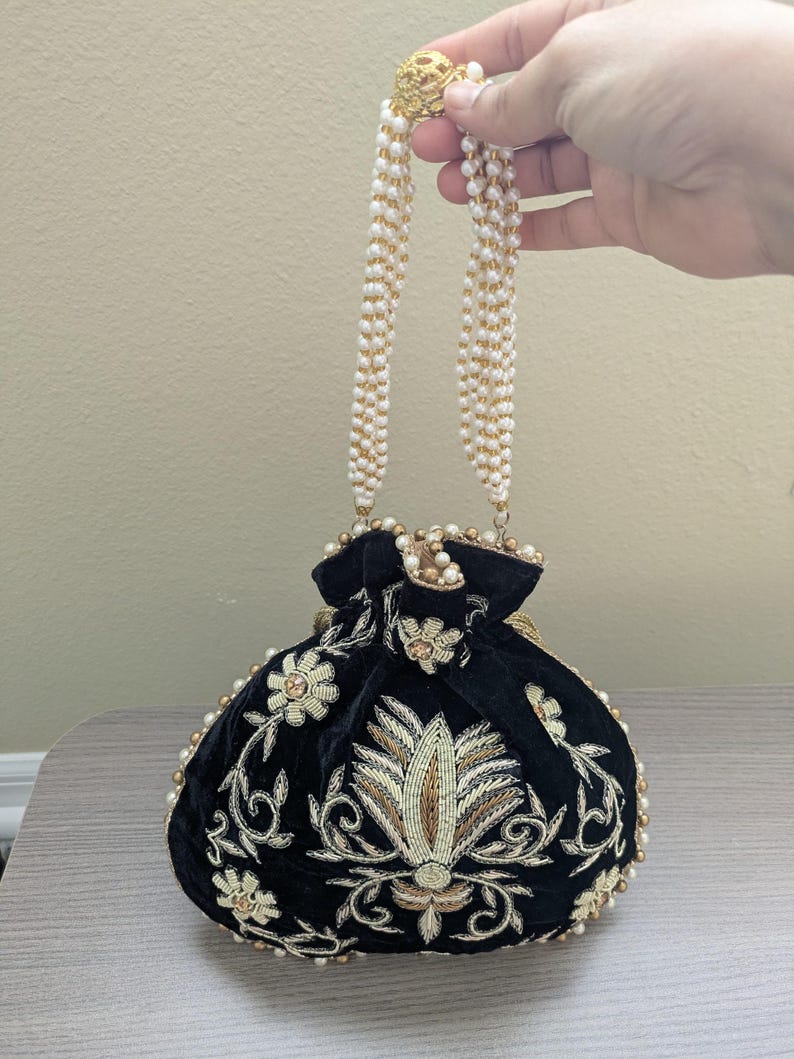 May include: Black velvet pouch with gold and cream floral embroidery. The bag features a beaded handle and a gold clasp. The pouch is adorned with pearls around the edges and on the handle.