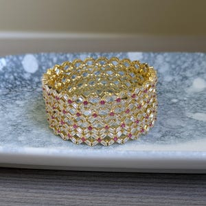 May include: A gold-toned bracelet with a geometric design. The bracelet features a repeating pattern of clear and red stones, creating a sparkling effect. The jewelry is displayed on a light-colored tray.