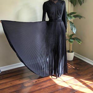 May include: A black, long-sleeved dress with a pleated skirt. The dress has a V-neck and a fitted bodice. The skirt is floor-length and flows outward. The dress is displayed on a mannequin in front of a houseplant.