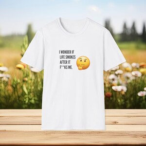 Op de afbeelding: Wit t-shirt met de tekst "I wonder if life smokes after it f**ks me" en een denkende emoji. Het shirt wordt getoond op een houten oppervlak met een vage achtergrond van bloemen en groen.