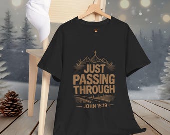 May include: Black t-shirt with the text "JUST PASSING THROUGH" in a brown font, featuring a mountain and cross design. The text "JOHN 15:19" is below the main text. A white baseball cap rests on a wooden chair.