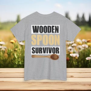 May include: A grey t-shirt featuring the text "WOODEN SPOON SURVIVOR" in black and gold, with a wooden spoon illustration. The shirt is shown on a wooden surface, with a blurred backdrop of flowers and foliage.