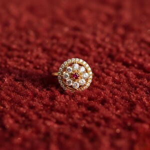 May include: A gold ring with a circular design, set with small white diamonds and a central red gemstone. The ring is on a red textured surface, creating a contrast of colors and textures. The ring's design is floral-inspired.