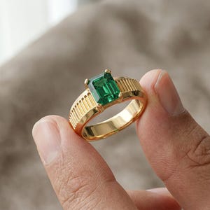 May include: A gold ring with a large, emerald-cut green gemstone. The ring has a gold band with vertical lines on the sides. The ring is held between two fingers.