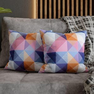 May include: Two square throw pillows with a geometric pattern in shades of pink, orange, blue, and purple rest on a gray sofa. A plaid blanket is draped over the back of the sofa. The pillows add a pop of color to the neutral-toned living space.