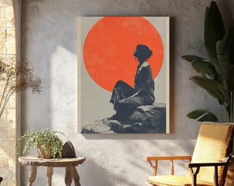 May include: A framed art print featuring a silhouette of a person seated on a rock, with a large orange circle in the background. The artwork is displayed in a room with a rustic aesthetic, including wooden beams and a small wooden table.
