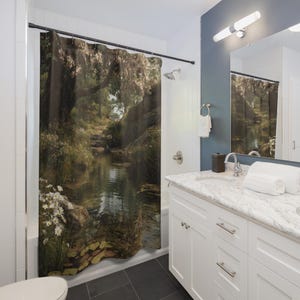 May include: A shower curtain featuring a serene landscape with a river flowing through a forest. The design includes trees, rocks, and flowers, creating a natural and calming scene. The curtain is hung in a bathroom with white cabinets and a marble countertop.
