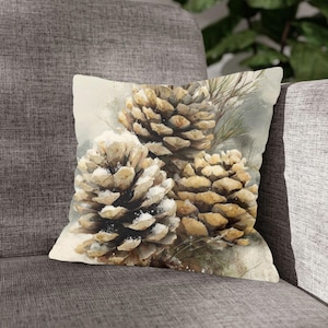 May include: A decorative throw pillow featuring a watercolor painting of three pine cones dusted with snow. The pine cones are shades of brown and tan, with a light gray and green background. The pillow is square and rests on a gray upholstered chair.