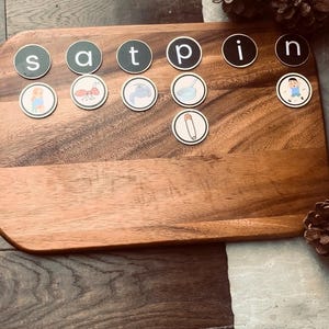 May include: Wooden board with magnetic letter tiles spelling words. The tiles are black with white letters and feature illustrations. Pine cones are in the background. Educational toy for children.