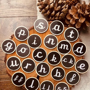 May include: A collection of small, round, black wooden discs with white cursive letters arranged on a wooden slice. The letters spell out words. The arrangement is surrounded by pine cones.