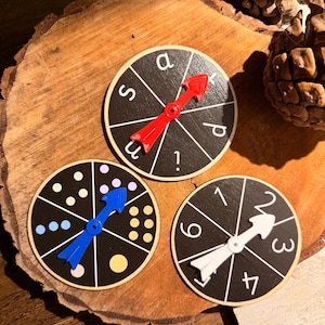 May include: Three wooden game spinners with black faces and white markings. Each spinner has a different coloured arrow and design. One has letters, one has dots, and the other has numbers. The spinners are on a wooden surface.