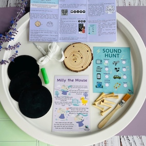 May include: A white tray displays educational resources. It features black circles, a wooden slice, a green crayon, a brush, and a card with "Milly the Mouse." There are also small wooden tees, a white cord, and cards with text.
