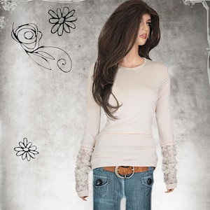 May include: A long-sleeved, cream-colored shirt with lace detailing on the cuffs. The shirt is fitted and has a round neckline. The model is wearing blue jeans and a brown belt.