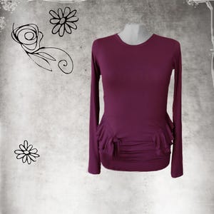 May include: A long-sleeved, plum-colored top with a round neckline and a gathered, ruffled detail at the waist. The top is displayed on a mannequin against a textured, gray background with floral doodles.